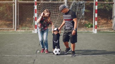 Full body of positive happy parents in casual clothes holding toddler son hands while walking together on football field on sunny day